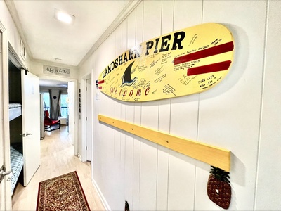 A hallway with a surfboard sign reading "Landshark Pier Welcome" on the wall. Nearby doors lead to other rooms. A rug lines the wooden floor. A small pineapple decoration is on the wall.