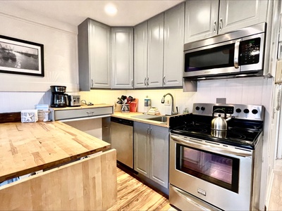 Modern kitchen with stainless steel appliances, light gray cabinets, a wooden countertop, and a coffee maker on the counter. A framed black-and-white photo hangs on the wall.