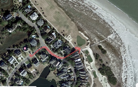 Aerial view of a coastal residential area with a road highlighted in red, houses, trees, a golf course, and adjacent beach and ocean visible.