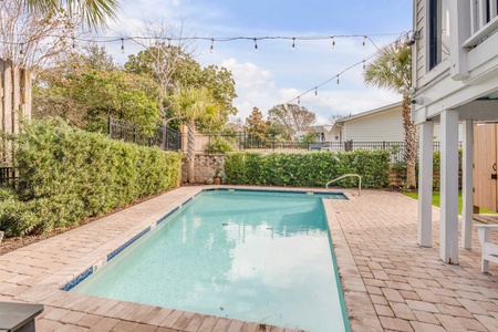 A rectangular outdoor pool with clear water surrounded by brick paving, greenery, and string lights overhead.