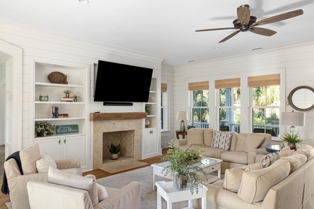 Living room with beige furniture, a wall-mounted TV above the fireplace, built-in shelves, large windows, ceiling fan, and a glass coffee table with plants.