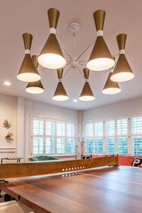 Modern room with a ping pong table and a large, gold chandelier. The room has white walls, decorative items on shelves, and large windows with shutters.