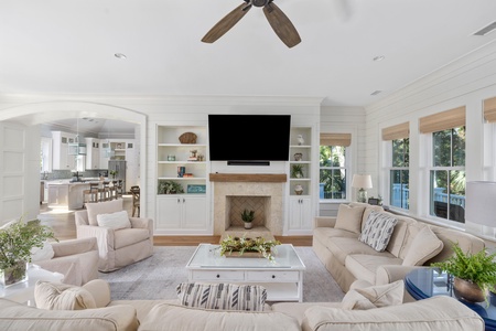 A bright living room with beige sofas, a coffee table, and a fireplace beneath a mounted TV. Shelves line the wall, and large windows provide natural light. The kitchen is visible in the background.