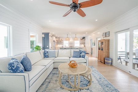 Living area with white sectional, cozy rug and round woven coffee table. Accents of blue keep the nautical theme fluid through the home. The kitchen is seen in the behind the sectional sofa.