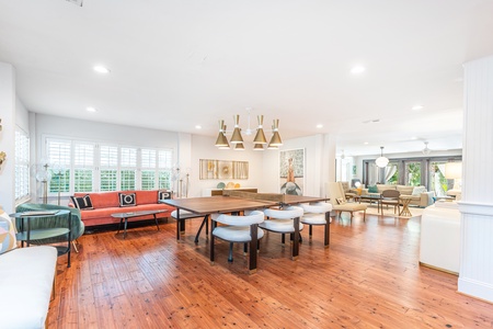 Dining room with modern retro decor. Large wood dinging table/ping pong table. Wood pine floors with white walls and ceiling. Large Brass chandelier and coral sofa with pillows I O P.