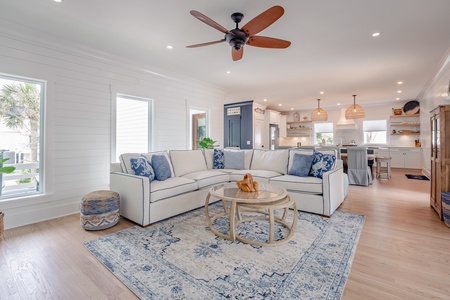 Living area with white sectional, cozy rug and round woven coffee table. Accents of blue keep the nautical theme fluid through the home. The kitchen is seen in the behind the sectional sofa.