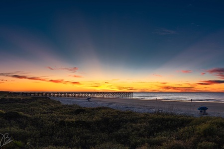 Sunset over a beach with a pier extending into the ocean, silhouetted against a sky with sun rays and scattered clouds.