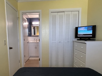 A small bedroom with a bed, dresser, wall-mounted TV, and a closet with shutter doors. An open door reveals a bathroom with a sink and bright lighting.