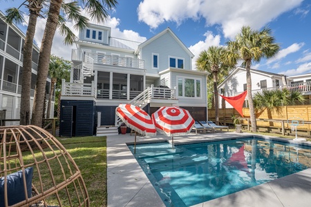 Two Red and White stiped pool unbrellas provide shade on the stairs of the in ground pool.  Four chaise lounge chairs around the pool. Stairs up to the deck and gas grill and a screen porch. A spiral staircase rises up to the second floor porch.