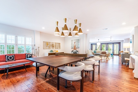 Dining room with modern retro decor. Large wood dinging table/ping pong table. Wood pine floors with white walls and ceiling. Large Brass chandelier.