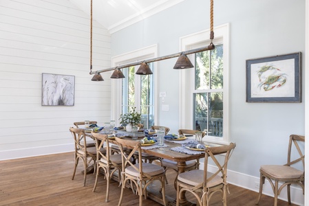 A dining room with a wooden table set for eight, pendant lights above, art on pale blue walls, and large windows.