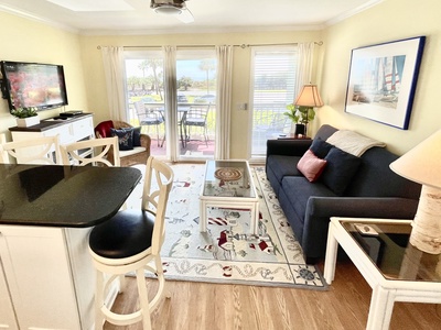 A bright living room with a navy sofa, a glass-top coffee table, a patterned rug, a wall-mounted TV, and bar stools by a kitchen counter. Sliding glass doors open to a patio with a view.