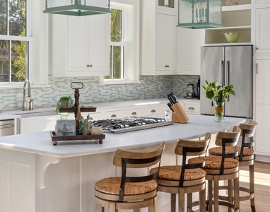 A bright kitchen with a central island, gas stove, wooden stools, and stainless steel fridge. Light blue tile backsplash and large windows enhance the airy atmosphere.