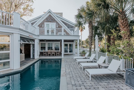 Poolside view of the home facing the back of the home. Four white chaise lounge sunning chairs on the right with umbrella. An exterior dining table under a cover by the kitchen.