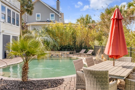 Backyard with a pool, palm tree, patio table with a red umbrella, and wicker chairs. Two lounge chairs by the pool. House and greenery in the background under a blue sky.