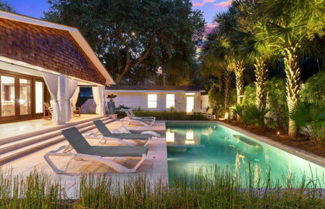 A backyard with a pool surrounded by lounge chairs and palm trees at twilight. A house with large windows and curtains is in the background.