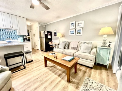 A cozy living room with a gray sofa, wooden coffee table, and side tables with lamps. A kitchen is visible with a fridge, cabinets, and a trash can. Photos are mounted on the wall.