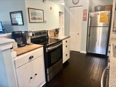 A modern kitchen with a stainless steel refrigerator, stove, and white cabinetry. A coffee maker sits on the counter beside the stove. Dark wood flooring contrasts with light-colored walls.