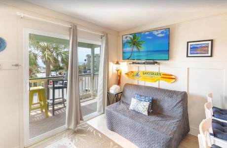 Cozy living room with a sofa, surfboard wall decor, and a large TV displaying a beach scene. Balcony view through a glass door reveals palm trees and outdoor seating.