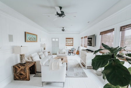 Bright living room with white walls and furniture, two ceiling fans, a wall-mounted TV, a guitar, and a large potted plant by the window.