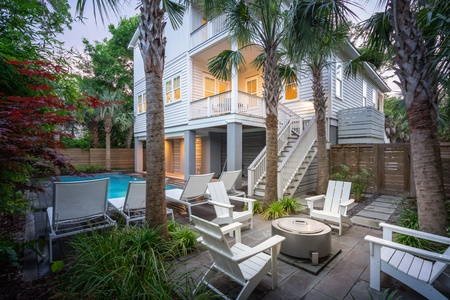 Outdoor area with white chairs around a fire pit, a pool, and palm trees. A two-story house with a balcony and lit windows is in the background.