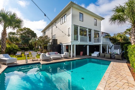 A modern two-story house with a backyard pool, surrounded by patio furniture and palm trees, under a clear blue sky.