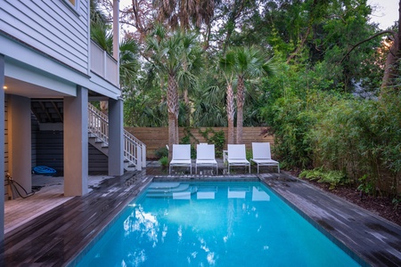 A backyard with a rectangular pool, surrounded by wooden decking. Four white lounge chairs are lined up beside the pool, bordered by a fence and lush greenery.