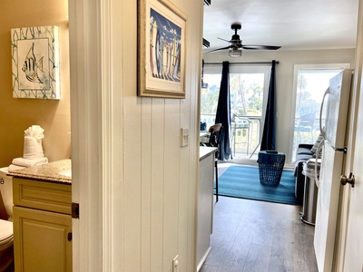 View from a hallway showing a bathroom on the left and a living area with balcony access. The space is decorated with nautical-themed artwork and has a ceiling fan and natural lighting.