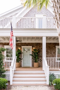 Front view of a house with a porch, American flag, shingle siding, and two potted plants by the mint green door. White railings and a palm tree are also visible.