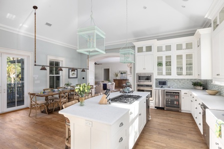 Spacious kitchen with white cabinets, island, and stainless steel appliances. Dining area with wooden table and chairs. Bright natural light from large windows.