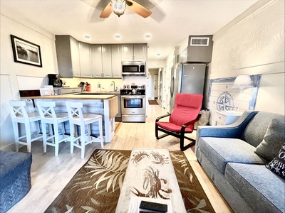 Cozy living room and kitchen with a red chair, gray sofa, rug, and bar stools at a counter. Kitchen has stainless steel appliances and wood cabinets. Ceiling fan above.