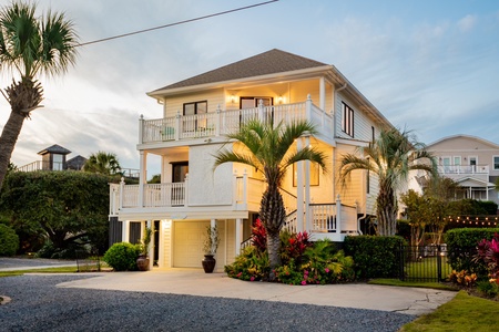 A two-story house with a balcony, surrounded by palm trees and a garden. The exterior is lit with warm lights during dusk.