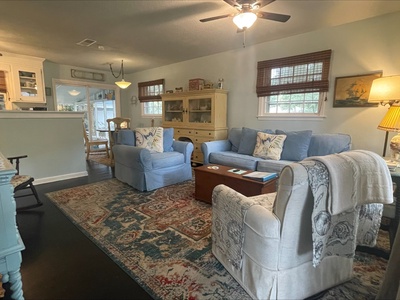 A cozy living room with blue and beige chairs, a rug, a wooden coffee table, wall art, and a ceiling fan. A kitchen is visible in the background.