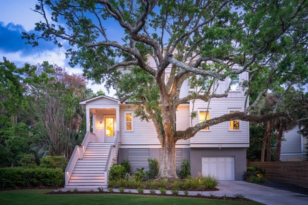 Two-story white house with large tree in front, illuminated windows, and a garage, set against a backdrop of greenery and evening sky.