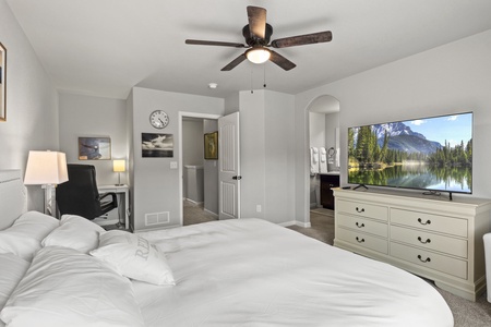 A bedroom features a white bed, ceiling fan, and television on a dresser showing a nature scene. A desk and chair are visible in the corner, and an open door leads to a bathroom.