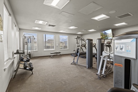 A small, well-lit gym room with exercise equipment, including treadmills, a weight rack with dumbbells, a bench, and various workout machines. Large windows provide natural light.