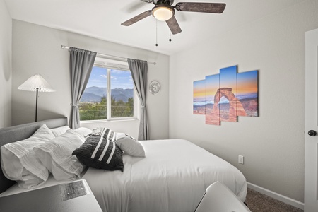A bedroom with a bed, pillows, and a gray headboard. A ceiling fan is above, and a window shows a mountain view with gray curtains. A multi-panel landscape artwork is on the wall.