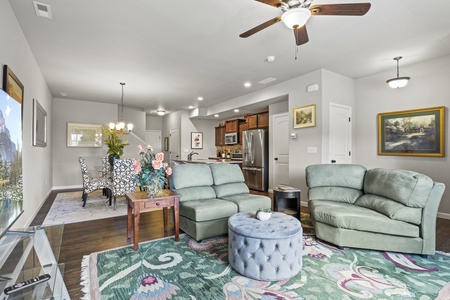 Modern living room with light gray walls and dark hardwood floors, furnished with green seating, a floral-patterned rug, and various wall art. The dining area and kitchen are visible in the background.