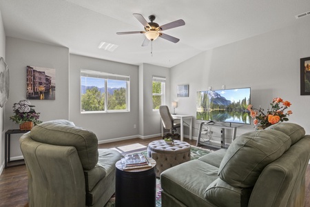 A living room with green armchairs, a small round ottoman, and a TV displaying nature scenery. There's a desk with a chair by the window, and plants decorate the room. Ceiling fan and light overhead.