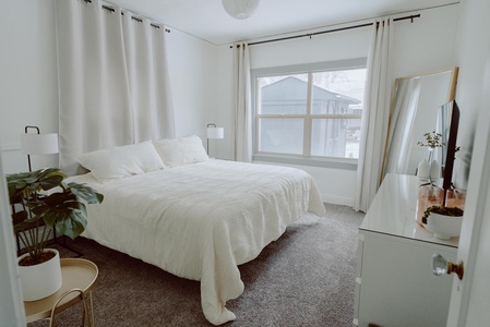 A bedroom with a large bed, white bedding, two bedside lamps, a large window, a plant, and a dresser with a mirror and decor. The room has light walls, curtains, and a carpeted floor.