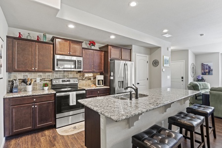 Modern kitchen with dark wooden cabinets, stainless steel appliances, granite countertops, and a large island with three leather stools. Open layout with a living area in the background.