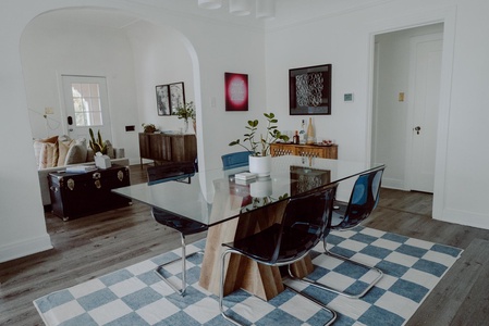 A dining room with a glass table, six blue chairs, a checkered rug, and various decor pieces. In the background is a living area with a couch and a coffee table. Walls are painted white.