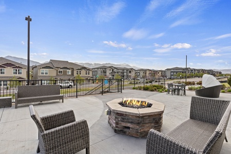 Outdoor patio with wicker chairs around a fire pit, set against a backdrop of townhouses and a clear blue sky.