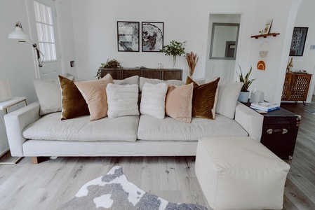 A living room with a beige sofa adorned with multiple cushions, situated on a wooden floor. The room is decorated with plants, wall art, and a cowhide rug. A beige ottoman is in front.