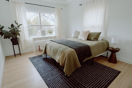 A tidy bedroom with a bed covered in green bedding, a rug, a desk with items under a window, two nightstands with lamps, and a potted plant near the window.