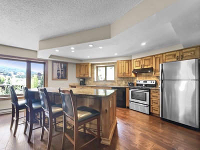 A modern kitchen with stainless steel appliances, wooden cabinets, a large central island with four chairs, and windows providing natural light and a scenic view.