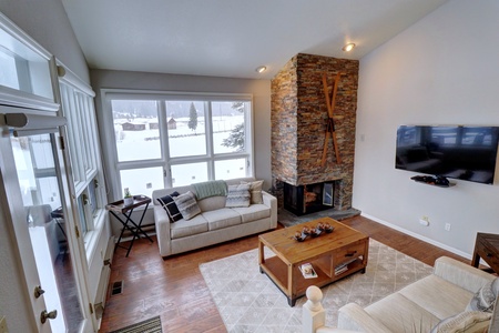 A cozy living room with a large window showing a snowy outdoor view. The room has a stone fireplace, a mounted TV, two sofas, a coffee table, and a wooden floor.