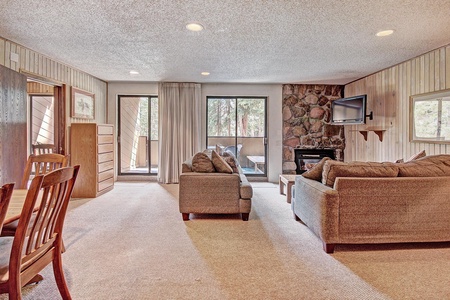 A living room features two sofas, a wall-mounted TV, a stone fireplace, and a glass sliding door leading to a balcony. The room has wood-paneled walls and a dining table with chairs on one side.