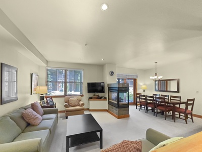 A well-lit living and dining area with beige walls. The space includes a sofa, armchair, TV, coffee table, and a dining table with eight chairs. A double-sided fireplace separates the two spaces.