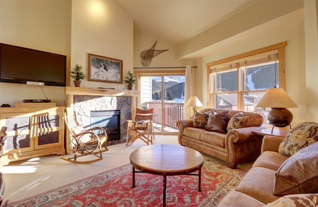 A cozy living room with a fireplace, mounted TV, brown leather sofas, wooden furniture, and large windows letting in natural light. A red patterned rug covers the floor. A wall-mounted decor seen.